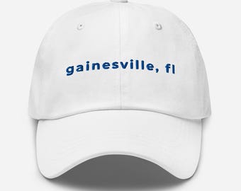 Könnte beinhalten: Eine weiße Baseballkappe mit dem gestickten Text "gainesville, fl" in Blau. Die Kappe hat einen gebogenen Schirm und ein sechsteiliges Design, geeignet für Freizeit.