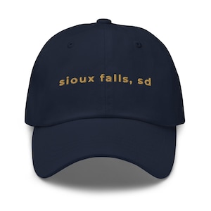Peut inclure: Casquette de baseball bleu marine avec le texte brodé "sioux falls, sd" en or. La casquette a une visière incurvée et un bouton sur le dessus.