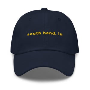 Op de afbeelding: Marineblauwe baseballcap met de geborduurde tekst "south bend, in" in geel. De cap heeft een gebogen klep en een knoop aan de bovenkant. Een klassiek, casual accessoire.