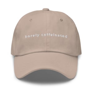May include: A beige baseball cap with the embroidered text "barely caffeinated" in white. The cap has a curved brim and a fabric strap closure on the back. The hat is a neutral color and suitable for everyday wear.