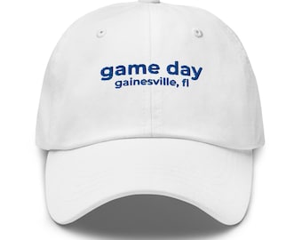 Könnte beinhalten: Weißes Baseball-Cap mit blauem Schriftzug "game day" und darunter "gainesville, fl". Die Kappe hat einen gebogenen Schirm und einen Knopf oben. Ein lässiges Accessoire.