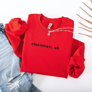 Puede incluir: Una sudadera roja con el texto "cincinnati, oh" en negro. La sudadera se combina con unos vaqueros azules claros rasgados y un collar plateado. Una hoja de palma seca está en el fondo.