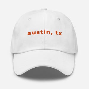 May include: White baseball cap with the embroidered text "austin, tx" in orange. The cap has a curved brim and an adjustable strap at the back. A casual accessory for everyday wear.