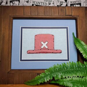 May include: A framed cross-stitch of a red and white straw hat with a white X on the crown. The hat is on a blue background.