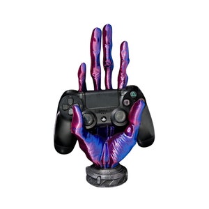 May include: A black PlayStation controller held in a purple and blue iridescent skeletal hand, which is a stand for the controller.