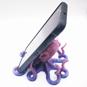 Op de afbeelding: Een glinsterende octopus telefoonstandaard in paars en blauw die een zwarte smartphone vasthoudt.