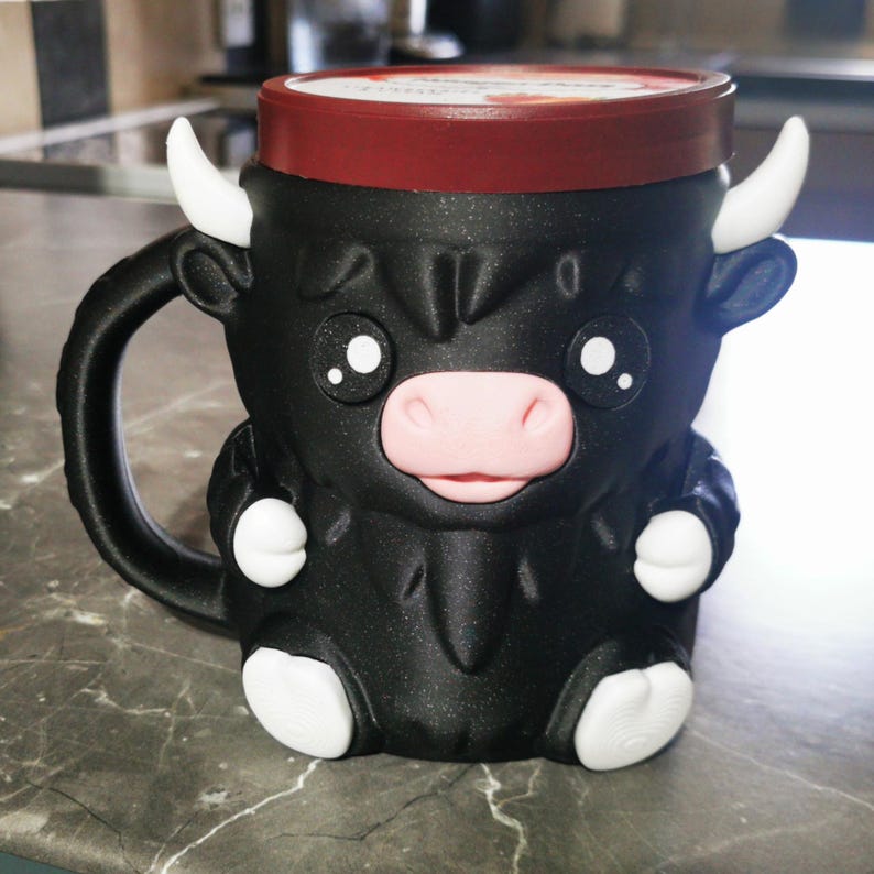 May include: A black ceramic mug shaped like a cartoon bull with white horns, hooves, and a pink nose. The mug has a red lid and a black handle. The mug is sitting on a gray countertop.
