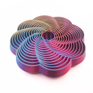 Puede incluir: Objeto iridiscente en forma de flor, con un diseño en capas. Los colores del objeto cambian de azul a morado y rosa, con un toque de amarillo. El diseño está hecho de capas curvas concéntricas.