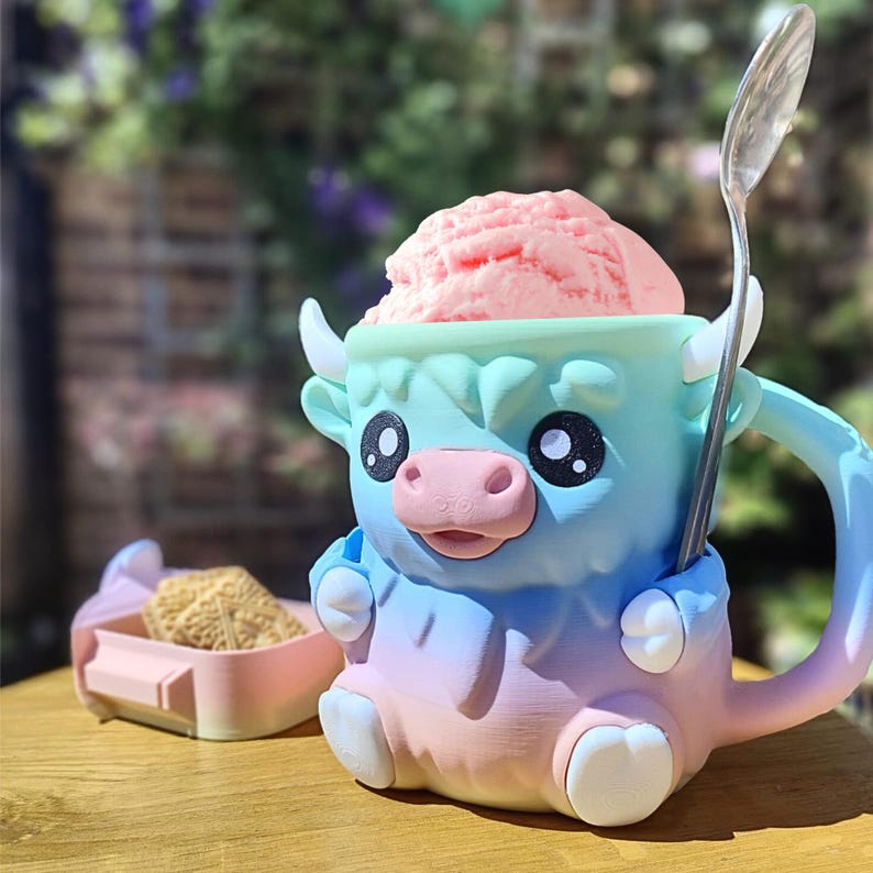May include: A whimsical, colorful mug shaped like a cartoon yak, filled with pink ice cream and a silver spoon. The mug is a gradient of blue, green, and pink. A small pink and blue container with a snack sits beside the mug.