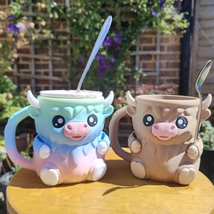 May include: Two ceramic Highland cow mugs. One is pastel blue, pink, and yellow with a lid and spoon. The other is tan with a spoon. Both mugs have horns, pink noses, and large eyes. The mugs are on a wooden surface.