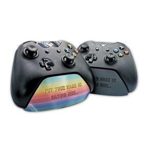 May include: Two black Xbox controllers with a rainbow-coloured design on the bottom of one controller. The text "PUT YOUR NAME OR SAYING HERE..." is engraved on the bottom of the controller.
