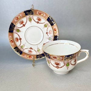 Puede incluir: Un juego de taza y platillo de té vintage con un patrón floral en tonos rojos, verdes, azules y dorados. La taza y el platillo tienen un fondo blanco con un borde dorado.