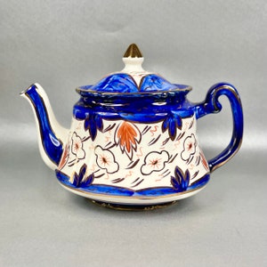 Op de afbeelding: Een blauwe en witte theepot met een bloemenmotief en gouden accenten. De theepot heeft een rond lichaam en een tuit aan de linkerkant. De handgreep bevindt zich aan de rechterkant.