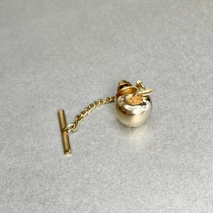 May include: A gold tie tack with a silver apple design. The apple has a small leaf on top.