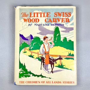 May include: A vintage book cover with a colorful illustration of a young boy in a red and white shirt and a blue hat, holding a horn. The book title is "The Little Swiss Wood Carver" by Madeline Brandeis. The book is part of "The Children of All Lands Stories" series.