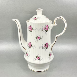 Puede incluir: Cafetera de porcelana blanca con ribete dorado y un patrón floral de rosas rosas y hojas verdes.