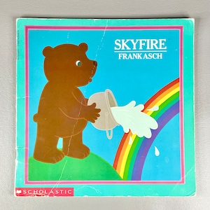 May include: A brown bear pours water from a silver bucket onto a rainbow. The book cover is blue with the title "Skyfire" and the author "Frank Asch" in white text. The book is published by Scholastic.