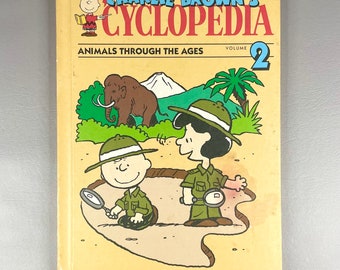 Könnte beinhalten: Ein gelber Buchumschlag mit dem Titel "Charlie Brown's Cyclopedia, Band 2: Tiere durch die Zeiten" und einer Illustration von Charlie Brown und Snoopy, die ein Mammut betrachten.