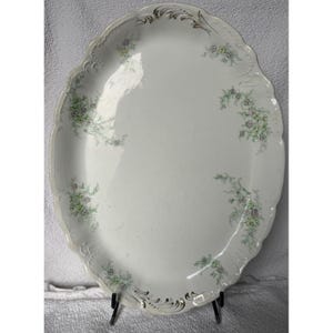 May include: A white oval serving platter with a scalloped edge and a floral design in green and pink. The platter has a gold trim around the edge.