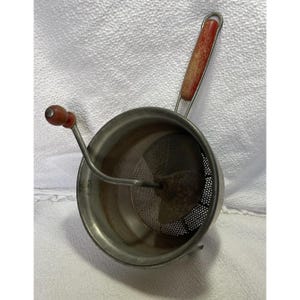May include: A vintage food mill with a stainless steel bowl and perforated disc. It features a wooden handle with a red finish and a crank with a wooden knob. This kitchen tool is designed for pureeing fruits and vegetables.