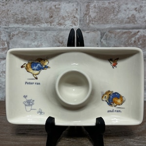 Puede incluir: Un huevo de cerámica blanco con un diseño de Peter Rabbit. El huevo tiene un plato pequeño a cada lado. Un lado presenta a Peter Rabbit corriendo con el texto "Peter ran". El otro lado presenta a Peter Rabbit corriendo con el texto "and ran."