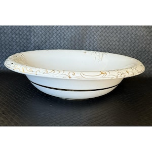 Pode incluir: Uma tigela de cerâmica branca com uma borda decorativa dourada e uma faixa preta. A tigela oval é provavelmente para servir ou exibir. Os detalhes dourados adicionam elegância. Medidas em centímetros.