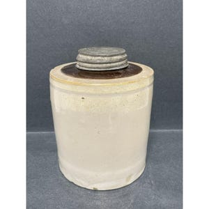 May include: A vintage stoneware crock with a metal lid. The cylindrical container is off-white with a brown rim and a metal lid. The crock has a rustic, aged appearance, suggesting it is an antique or a piece of farmhouse decor.