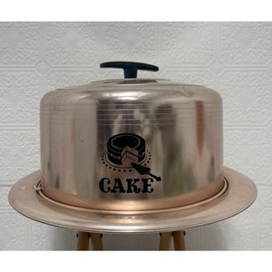 May include: A vintage, rose gold cake dome with a black handle and a black graphic of a cake slice with the word "CAKE" underneath.