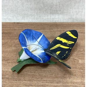 Puede incluir: Figura decorativa con una flor de gloria de la mañana azul y blanca y una mariposa negra y amarilla. La mariposa tiene rayas amarillas en sus alas. La flor y la mariposa descansan sobre una hoja verde.