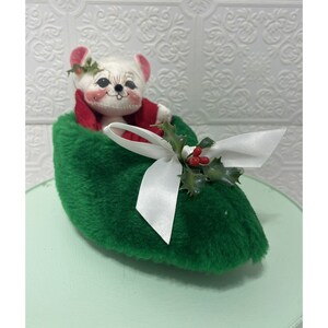 May include: A white mouse plush toy wearing a red hat and sitting in a green felt slipper with a white bow and holly sprig.