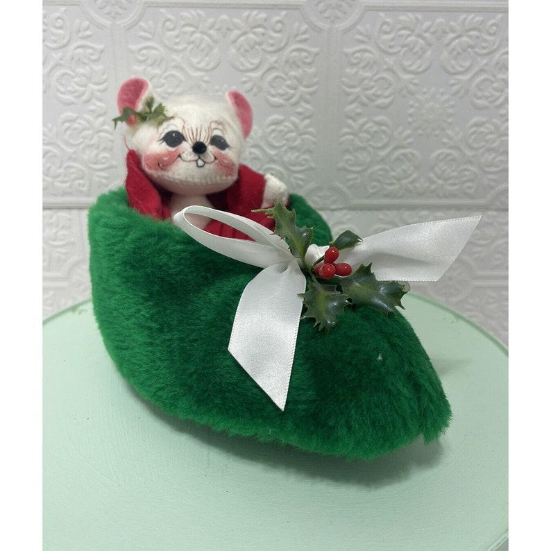 May include: A white mouse plush toy wearing a red hat and sitting in a green felt slipper with a white bow and holly sprig.