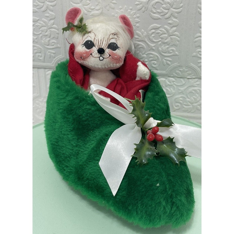 May include: A white mouse plush toy wearing a red hooded robe sits inside a green plush stocking with a white ribbon and holly sprig.