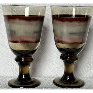 May include: Two vintage wine glasses with a unique design. The glasses feature a dark brown stem and base, transitioning to a gold band, and then a layered pattern of red, gray, and clear glass. The rims are clear.