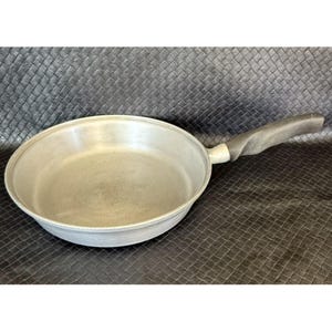May include: A silver-colored frying pan with a black, twisted handle. The pan is round with a wide, shallow cooking surface. The handle is ergonomically designed for a comfortable grip. The pan is likely made of aluminum or a similar metal.