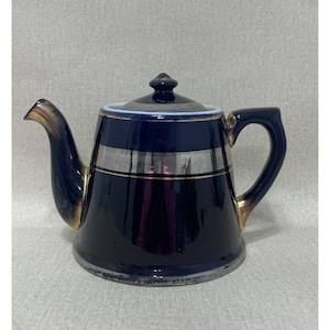 May include: A navy blue teapot with a silver band and gold accents. The teapot has a lid with a small knob and a curved handle. The spout and handle are trimmed in gold. The teapot is likely made of ceramic.