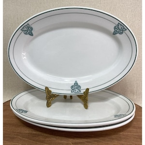 May include: White oval serving platters with a teal green border and decorative corner accents. The platters are stacked on a gold-coloured metal stand. The top platter is in focus, with two more platters stacked below.