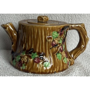 May include: A brown ceramic teapot shaped like a tree stump with green and red floral details.