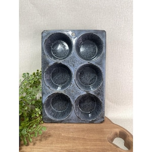 Könnte beinhalten: Eine Vintage-Muffinform für sechs Tassen mit gesprenkelter blau-grauer Emaille-Oberfläche. Die rechteckige Form hat sechs runde Tassen und ein kleines Loch oben zum Aufhängen. Die Form steht auf einer Holzoberfläche.