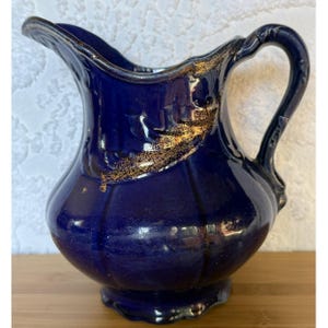 May include: A navy blue ceramic pitcher with a gold-colored accent. The pitcher has a curved handle and a spout for pouring. The gold accent is applied in a decorative pattern along the rim and side of the pitcher. The pitcher is a decorative item.