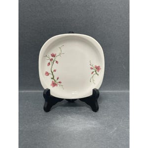 Puede incluir: Un plato cuadrado de cerámica blanca con un diseño floral con flores rosas y tallos verdes. El plato está sobre un soporte negro.