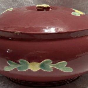 May include: A vintage red ceramic casserole dish with a lid. The dish has a floral design with green leaves and yellow flowers.