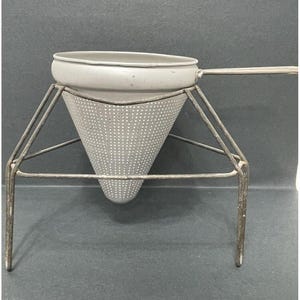 May include: A vintage metal strainer with a conical shape and a handle. The strainer is sitting on a metal stand with three legs.