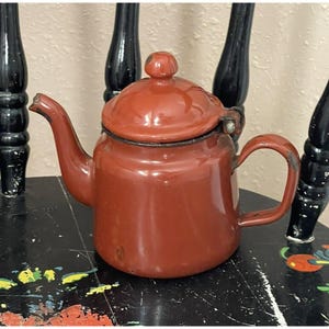 Peut inclure: Une petite théière en émail rouge avec un couvercle et une poignée. La théière est posée sur une surface noire et blanche.