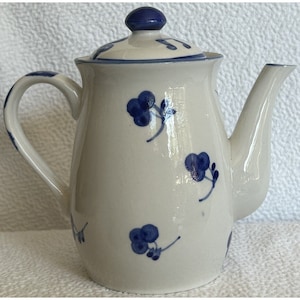 May include: A white ceramic teapot with a blue lid and blue floral designs. The teapot has a spout and a handle.