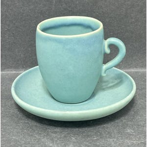 May include: A light blue ceramic teacup and saucer set. The cup has a rounded shape with a curved handle. The saucer is round and flat. The set has a matte finish and a slightly textured surface. The color is a soft, muted blue.