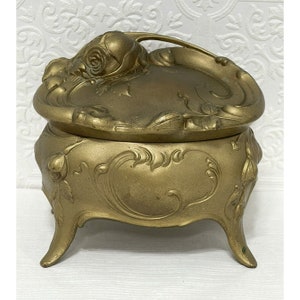 May include: A gold-toned metal trinket box with an ornate floral design. The box has a hinged lid and sits on three legs.