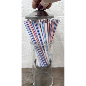 Puede incluir: Un dispensador de pajitas de vidrio transparente con tapa y base de acero inoxidable. El dispensador está lleno de pajitas a rayas rojas, blancas y azules. Una mano levanta la tapa. El dispensador está sobre una superficie de madera.