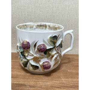 Peut inclure: Tasse à thé en céramique blanche avec une anse, ornée de motifs floraux en relief dans les tons de violet, vert et or. Le bord de la tasse est doré, et les détails floraux donnent un effet tridimensionnel à l'extérieur.