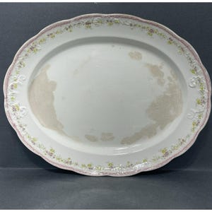 Peut inclure: Grand plat de service ovale blanc avec un bord festonné et une délicate bordure florale rose, jaune et violet. Le plat présente un aspect légèrement usé, avec une décoloration sur la surface.