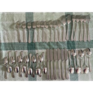 May include: A collection of vintage silverware, including forks, knives, and spoons, arranged on a green and white checkered tablecloth. The silverware is silver-colored and appears to be made of metal. The arrangement suggests a formal dining setting.